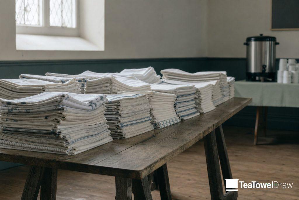 Folded tea towels stacked on a trestle table in a parish hall, ready to sell after a service.