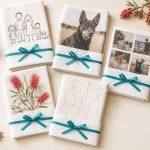 Five personalised tea towels half wrapped as Christmas presents, showing a child's drawing, a dog photo, family photos and a painting.